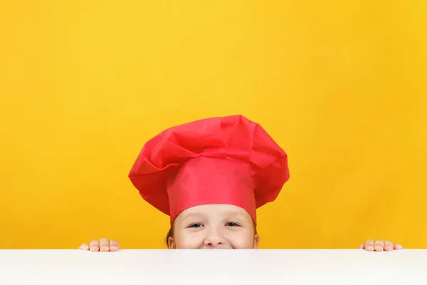 Zabawna mała kucharka na żółtym tle. Dziecko ukrywa się i patrzy spod stołu. — Zdjęcie stockowe