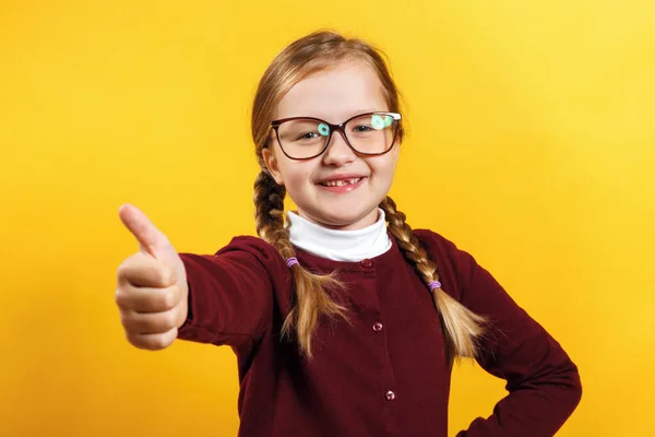 Student s brýlemi ukazuje palec nahoru. Malá dívka s copánky v vínové svetr na žlutém pozadí — Stock fotografie