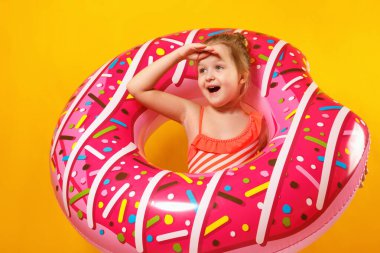 Mayo giymiş neşeli, güzel bir kızın portresi şişme donut halkasına bakıyor, sarı arka plan.