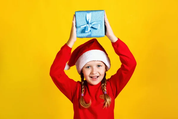 Felice bambino carino tiene una scatola sopra la testa. Bambina in cappello Babbo Natale con il Natale presente su sfondo giallo — Foto Stock