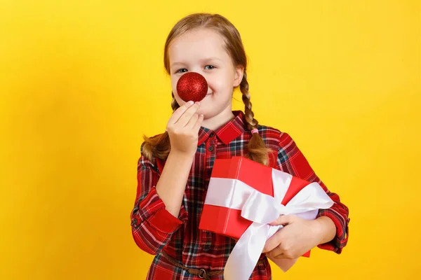 Vtipná šťastná holčička drží krabici s dárkem a dělá nos červeného vánočního míčku. Detailní záběr dítěte v červených šatech na žlutém pozadí — Stock fotografie