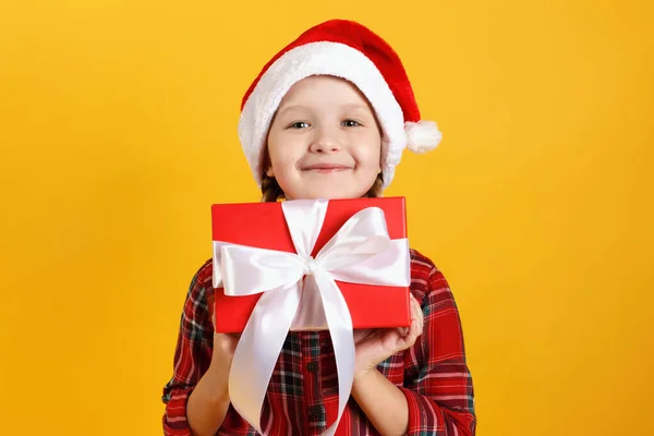 Счастливая маленькая девочка держит коробку с рождественским подарком. Ребенок в красном платье и шляпе санта на желтом фоне — стоковое фото