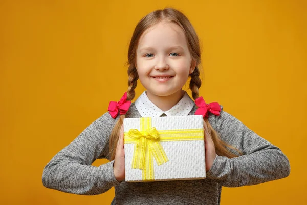 Ragazzina carina che tiene una scatola con un regalo in mano. Bambino con le trecce su sfondo giallo — Foto Stock