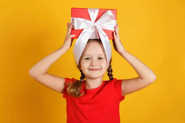 Allegra bambina felice tiene una scatola con un regalo in testa. Bambino in t-shirt rossa su sfondo giallo — Foto Stock