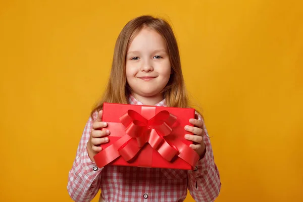 Bambino carino tiene una scatola rossa con un regalo. Bambina in camicia rosa su sfondo giallo — Foto Stock