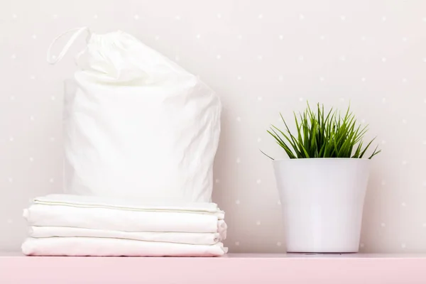 Une pile de literie propre, un sac de rangement, une plante domestique sur une table contre un mur léger — Photo