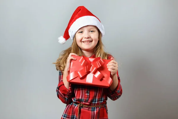 Concepto de Navidad y Año Nuevo. Linda niña encantadora en sombrero de santa y vestido rojo sobre un fondo gris. Un niño sostiene en su mano una caja con un regalo — Foto de Stock