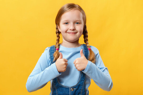 Cute happy little girl shows thumb up with two hands. A child with pigtails on a yellow background