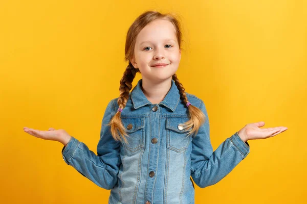 Roztomilá půvabná holčička ukazuje ruce na stranu. Dítě v džínové košili a s copánky na žlutém pozadí — Stock fotografie