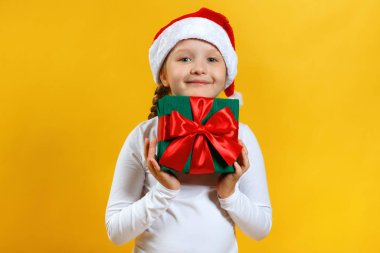 Noel Baba şapkalı mutlu çocuk Noel hediyesini tutuyor. Sarı arka planda beyaz tişörtlü küçük kız.