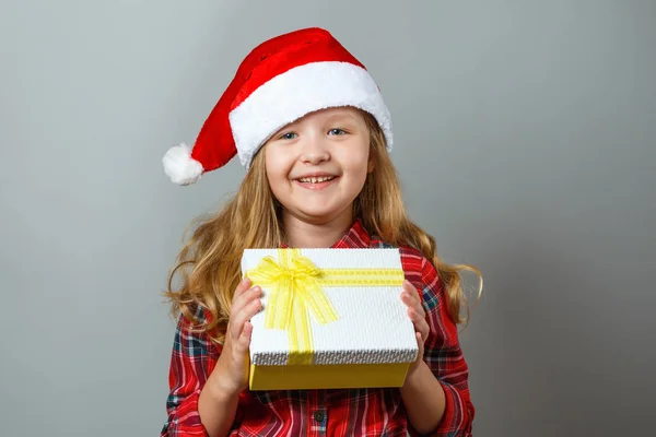 Natale e Capodanno concetto. Carino bambino allegro tiene una scatola con un regalo in mano. Affascinante bambina in cappello di Babbo Natale e vestito rosso su sfondo grigio — Foto Stock