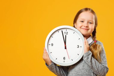 At kuyruklu küçük kız sarı arka planda büyük bir saat tutuyor. Eğitim, okul, zamanlama, öğrenme zamanı. Yakından bir portre. Boşluğu kopyala