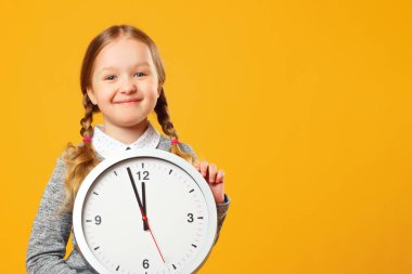 Sarı arka planda büyük bir saat tutan küçük bir kız. Eğitim, okul, teslim tarihi, öğrenme zamanı