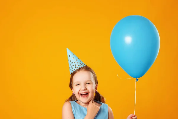 Cheerful little girl celebrates birthday. The child holds a blue ball and laughs. Closeup portrait on yellow background. Copy space — Stock Photo, Image