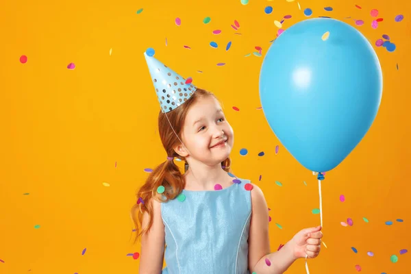 Cheerful little girl at a party. A child holds a blue ball in a rain of confetti. Closeup portrait on yellow background — Stock Photo, Image