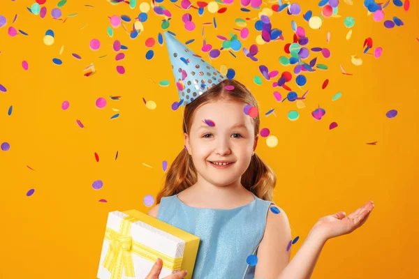 Cheerful little girl celebrates birthday. A child holds a box with a gift in the rain of confetti. Closeup portrait on yellow background — Stock Photo, Image