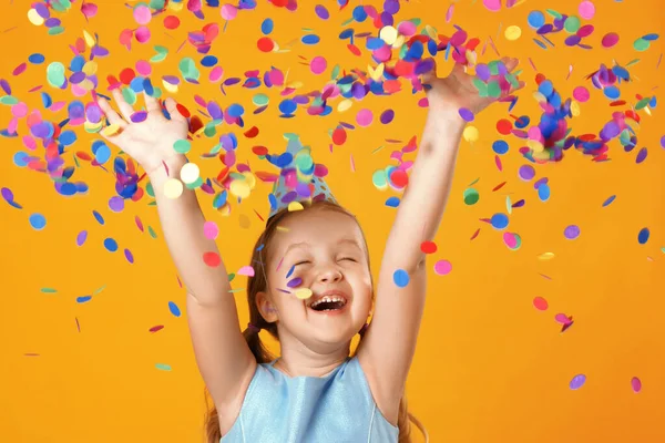 Vrolijk klein meisje viert verjaardag. Het kind staat met de ogen gesloten in de regen van confetti. Close-up portret op gele achtergrond — Stockfoto