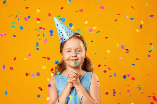 Cute little girl celebrates birthday. The child closed his eyes and makes a wish in the rain of confetti. Closeup portrait on yellow background — Stock Photo, Image
