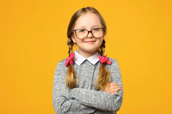 Detailní portrét legrační holčičky v brýlích na žlutém pozadí. Dětská školačka si založila ruce a dívá se do kamery — Stock fotografie