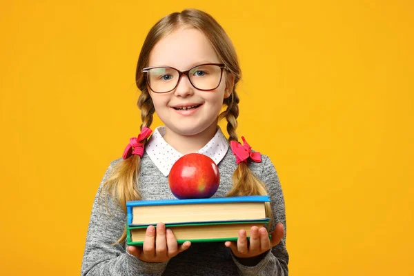 Ritratto di bambina allegra in occhiali su sfondo giallo. La studentessa tiene una pila di libri e una mela. Il concetto di istruzione. Ritorno a scuola — Foto Stock