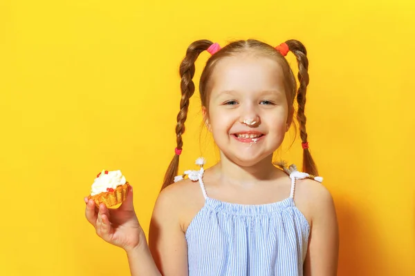 Detailní portrét veselé malé holčičky se sladkými zuby na žlutém pozadí. Dítě si rozmazalo nos smetanou a drželo v rukou dort. — Stock fotografie