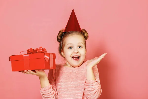 Closeup portrait of a little girl with wisps of hair over pink background. The child has a hat on his head and holds a box with a gift in his hands — Stock Photo, Image