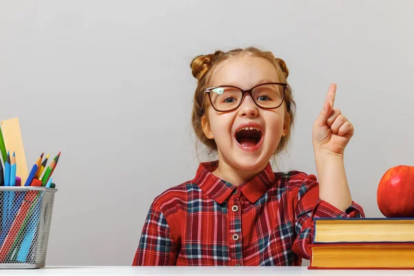 La ragazza prescolastica divertente in occhiali si siede al tavolo con libri e punta il dito su. Il concetto di istruzione. Fondo grigio — Foto Stock