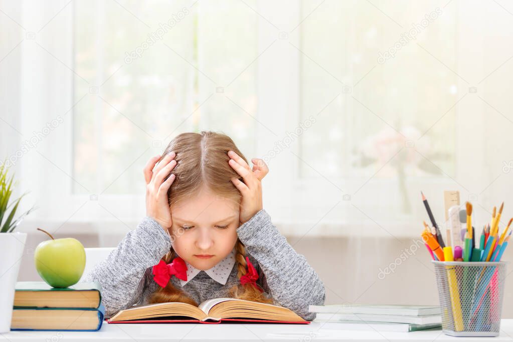 Little student sitting at the table and reading a book. The child learns and holds his head with two hands. The concept of education and school. Blurred background. Sunlight from the window