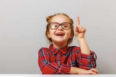 Okula dönelim. Gözlüklü komik küçük kız parmağını kaldırdı. Eğitim konsepti. Gri arkaplan