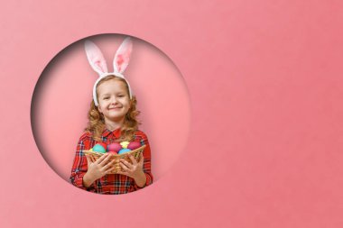 Paskalya yumurtası sepeti tutan tavşan kulaklı şirin bir kız çocuğu. Renkli pembe arka planda yuvarlak bir çember içindeki çocuk