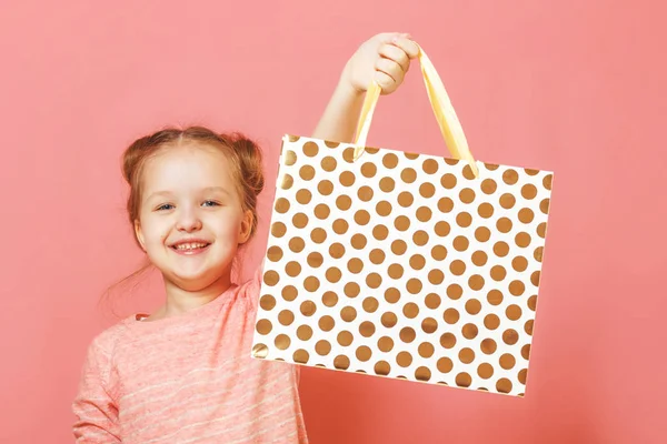 Nahaufnahme Porträt eines niedlichen kleinen Mädchens mit Haarbäckchen auf rosa Hintergrund. das Kind hob die Papiertüte hoch und freute sich über den Einkauf oder das Geschenk — Stockfoto