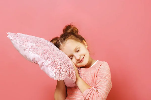 Люди и концепция здорового сна. Маленькая девочка обнимает мягкую подушку, закрывает глаза и позирует на фоне студии. Розовый фон — стоковое фото