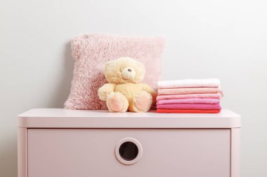 Bir yığın bebek giysisi ve pembe şifoniyerde bir oyuncak ayı. Gri duvar arkaplanı