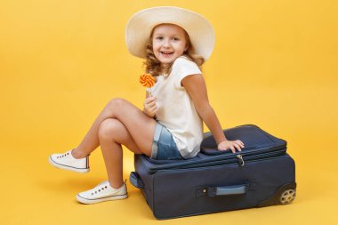 Sarışın bir kız çocuğu seyahat etmeyi hayal eder ve elinde şekerle bir bavula oturur. Sarı arkaplan