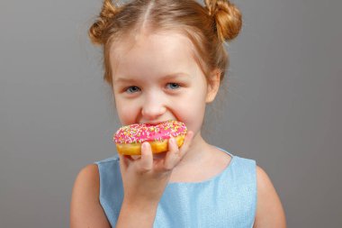 Mavi elbiseli şirin bir kız bebek lezzetli pembe bir donutu ısırıyor. Gri arkaplan, portre, stüdyo