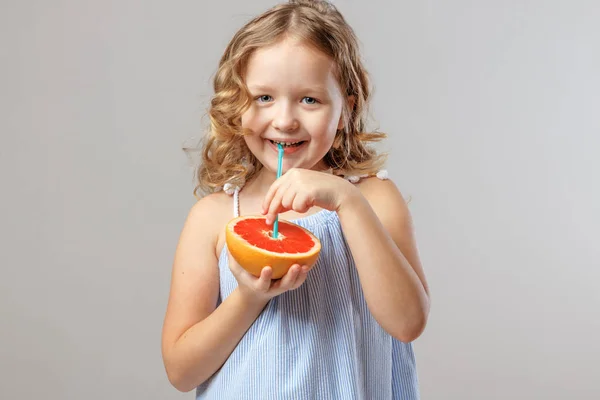Šťastné malé dítě drží půl grapefruitu a pije džus z tubulu. Zdravé jídlo. Šedé pozadí, studio — Stock fotografie