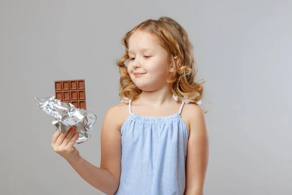 Encantadora niña niña rubia mira una barra de chocolate. Fondo gris, estudio, retrato — Foto de Stock