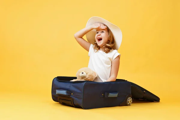 Mała blondynka dziewczynka marzy o podróży i siedzi w walizce z niedźwiedziem zabawki.Żółte tło — Zdjęcie stockowe