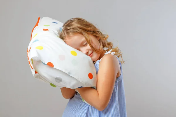 Ludzie i zdrowy sen. Mała dziewczynka przytula miękką poduszkę, zamyka oczy i pozuje na tle studia. Szare tło — Zdjęcie stockowe
