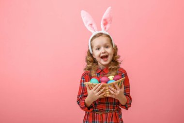 Renkli arka planda paskalya yumurtası sepeti tutan tavşan kulaklı şirin bir kız çocuğu.