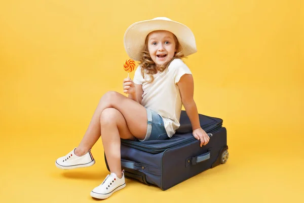 A little blonde kid girl dreams of traveling and sits on a suitcase with candy in her hand. Yellow background