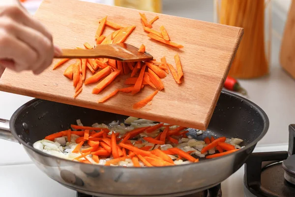 Домашняя кухня. Женщина с деревянной разделочной доской добавляет морковь в горячую сковороду с растительным маслом и луком. Крупный план — стоковое фото