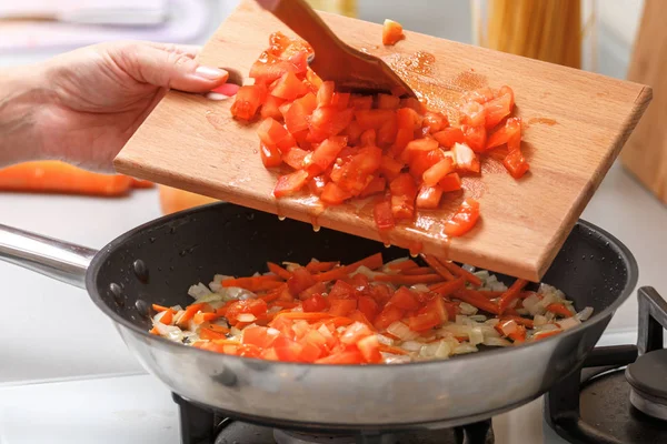Домашняя кухня. Женщина с деревянной разделочной доской добавляет помидоры в горячую сковороду с растительным маслом, луком и морковью. Крупный план — стоковое фото