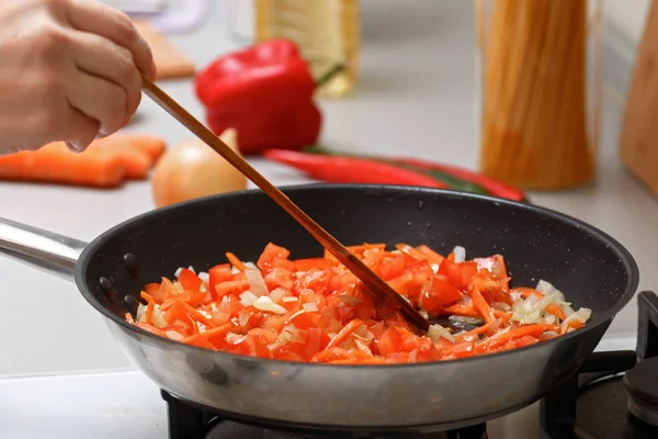 Домашняя кухня. Женщина перемешивает лук, морковь и помидоры в горячей сковороде с растительным маслом с деревянной лопаткой. Крупный план — стоковое фото