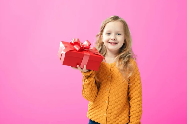 Glückliches kleines Mädchen in einem warmen Strickpullover, das eine Schachtel mit einem Geschenk hält, isoliert auf rosa Hintergrund. das Konzept des Schenken und Empfangens — Stockfoto