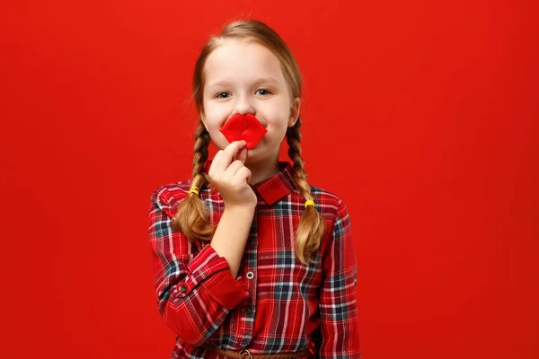 Funny cheerful little girl makes artificial fake lips on a red background. Child fooling around — Stock fotografie