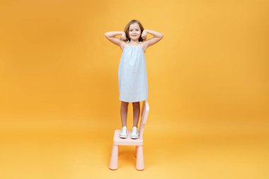 Küçük kız sandalyede duruyor ve güçlü kollarını gösteriyor.