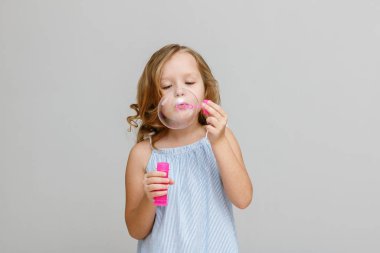 Gri arka planda mutlu, gülümseyen sarışın bir kızın portresi. Çocuk kabarcıkları üflüyor