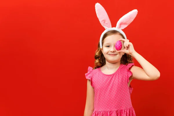 Schattig gelukkig klein meisje in konijnenoren houdt een paasei vast. Close-up portret van een kind op een rood gekleurde achtergrond — Stockfoto