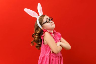 Mutlu Paskalyalar. Tavşan kulaklı, gözlüklü şirin bir kız kollarını kavuşturup yüzünü çevirmiş. Renkli kırmızı arka planda yakın plan bebek.
